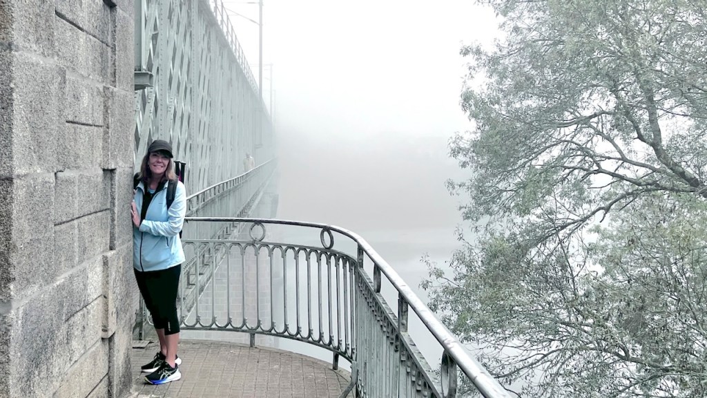 A woman in a light blue jacket and cap stands by a stone wall on a foggy bridge, surrounded by mist and greenery.