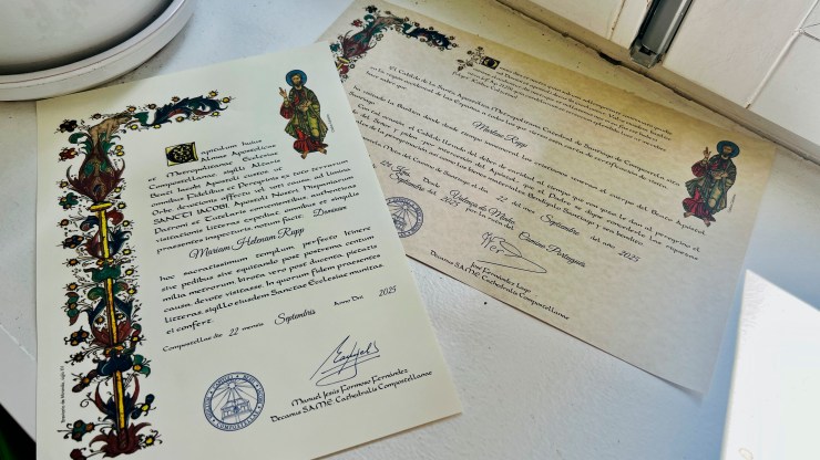 Two decorative certificates on a white surface, featuring colorful borders and illustrations of saints. The left certificate bears a name and date, while the right certificate has a different design and also includes a name and signatures.