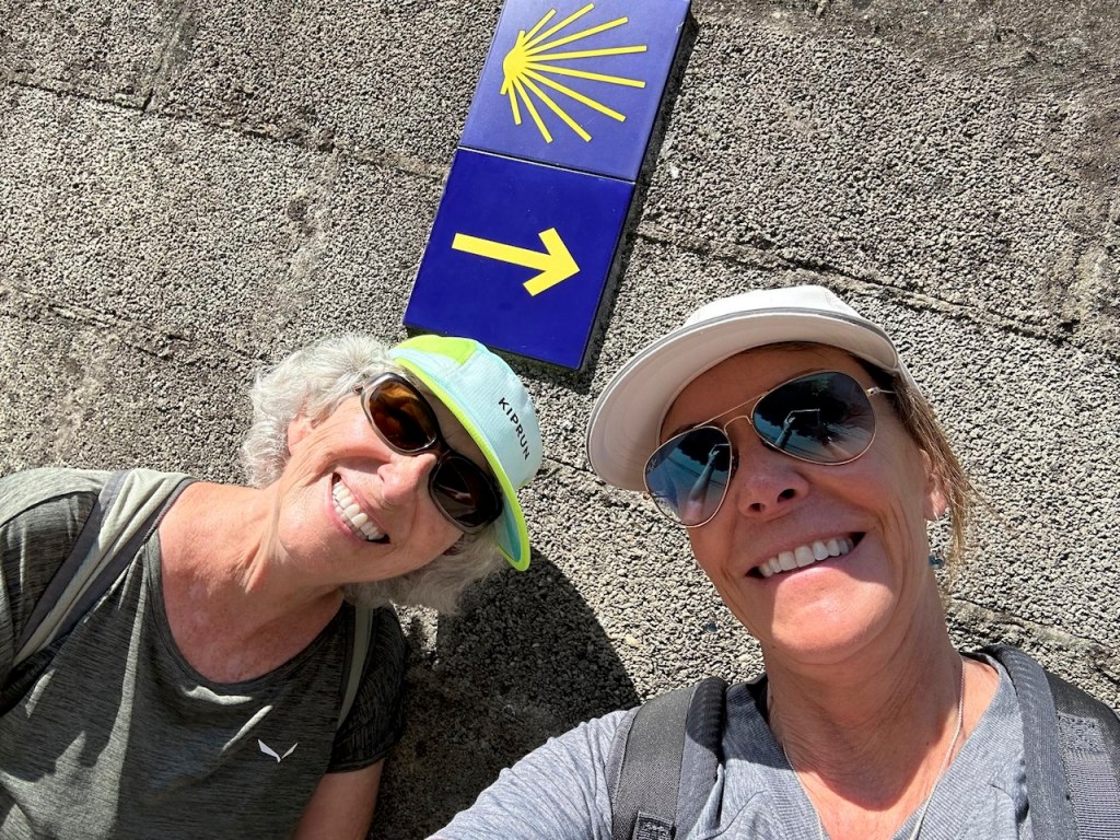 Two smiling women posing for a selfie in front of a directional sign with a yellow shell symbol, indicating a path.