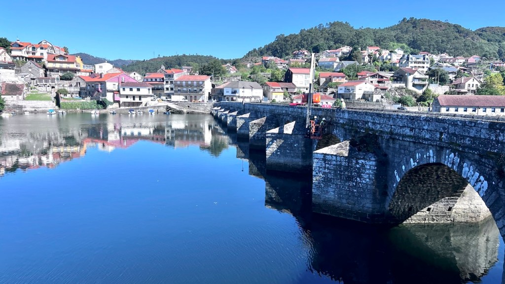A scenic view of a stone bridge over calm water, reflecting colorful buildings and a clear blue sky.