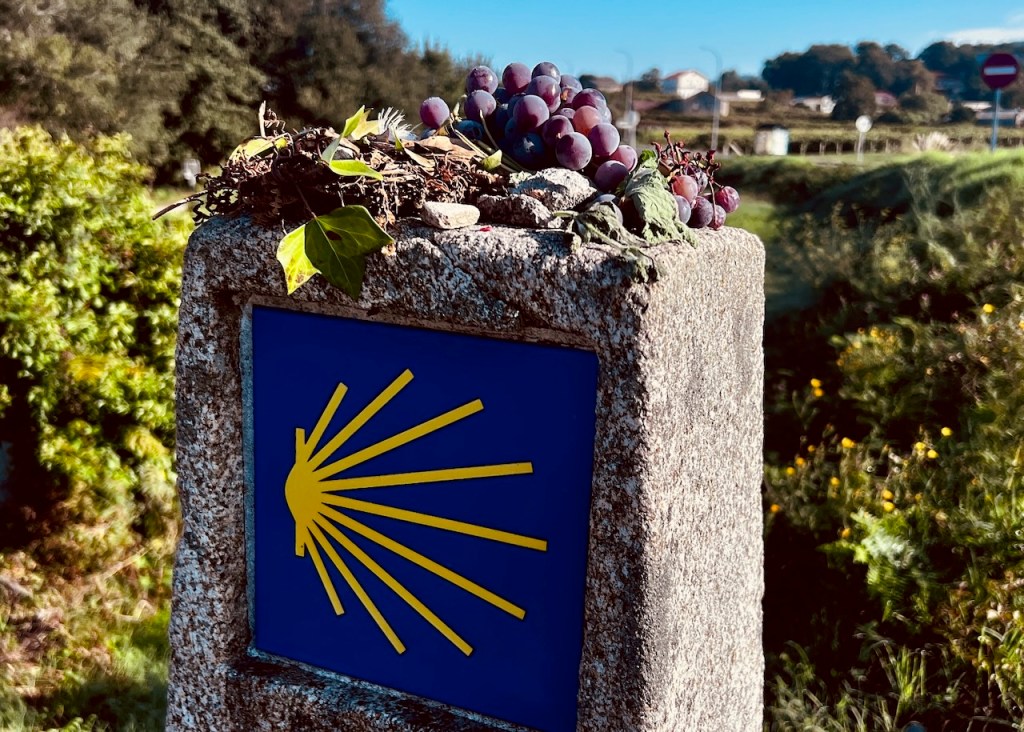 A stone marker with a blue painted background featuring a yellow sunburst symbol, adorned with grape vines and leaves on top, set against a lush green landscape.