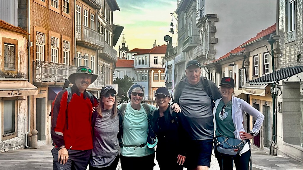 A group of six hikers posing together on a charming street lined with traditional buildings, smiling and wearing outdoor gear, with a sunset backdrop.
