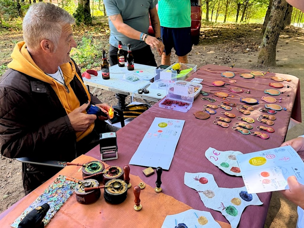 A man working at a table in an outdoor setting, crafting jewelry. There are various colorful pendants displayed on the table, along with tools and materials for making them. Several people are gathered around, and drinks are visible in the background.