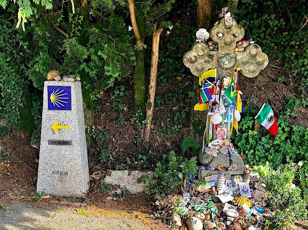 A Camino de Santiago milestone indicating 'Km 96.400' with an arrow pointing left, surrounded by greenery. Next to it, there is a decorative stone cross adorned with various items including flags, photographs, and mementos left by pilgrims.
