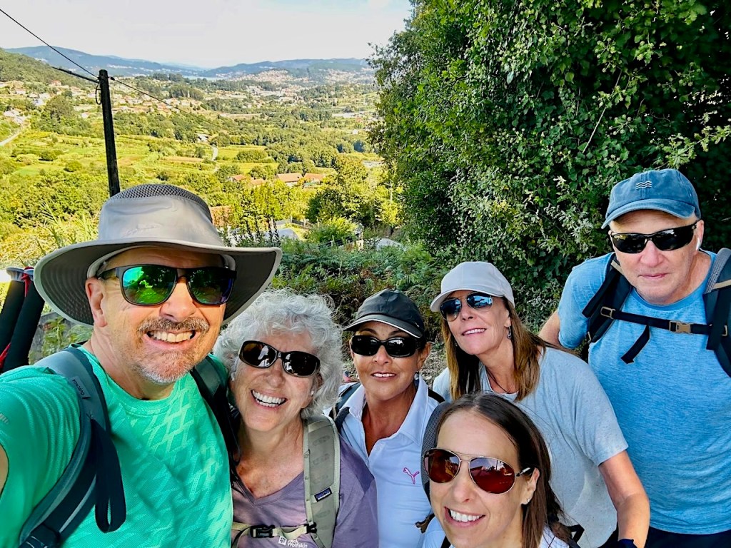 Group selfie of six hikers smiling on a trail with a scenic landscape in the background.