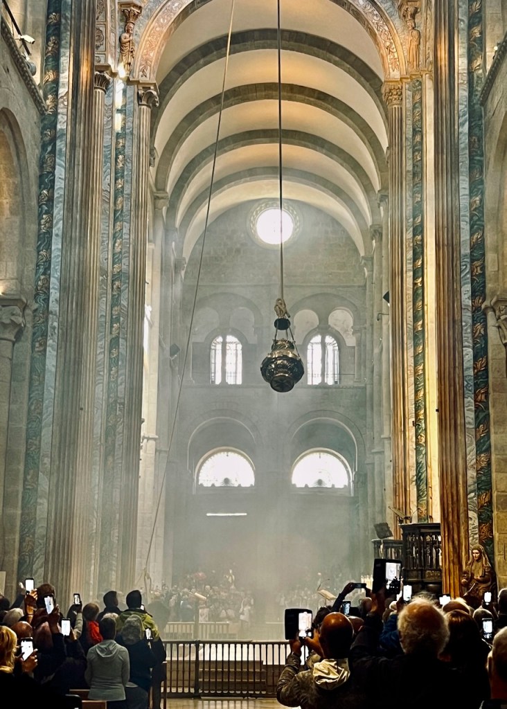 Interior of a cathedral with high arches and a large round window, filled with light and smoke. A large incense burner is suspended from the ceiling, while a crowd of people, some holding smartphones, watches an event.