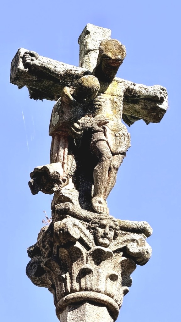 A weathered stone crucifix featuring a figure of Jesus on the cross, with intricate carvings and moss growth, against a clear blue sky.