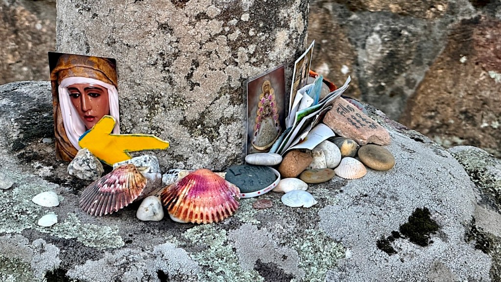 A small shrine featuring a colorful image of a figure, surrounded by shells, stones, and various postcards on a textured stone surface.
