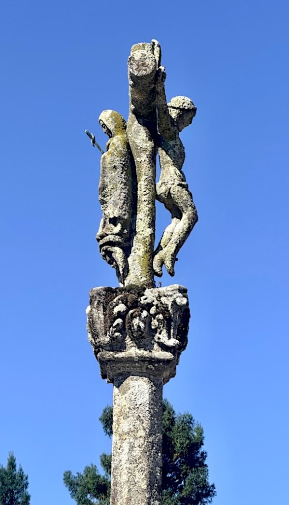 A weathered stone sculpture depicting two figures, one climbing a cross, atop a column with intricate carvings, against a clear blue sky.