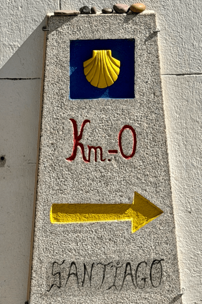 Milestone with a yellow shell emblem, indicating 'Km 0' and pointing towards Santiago with a yellow arrow.