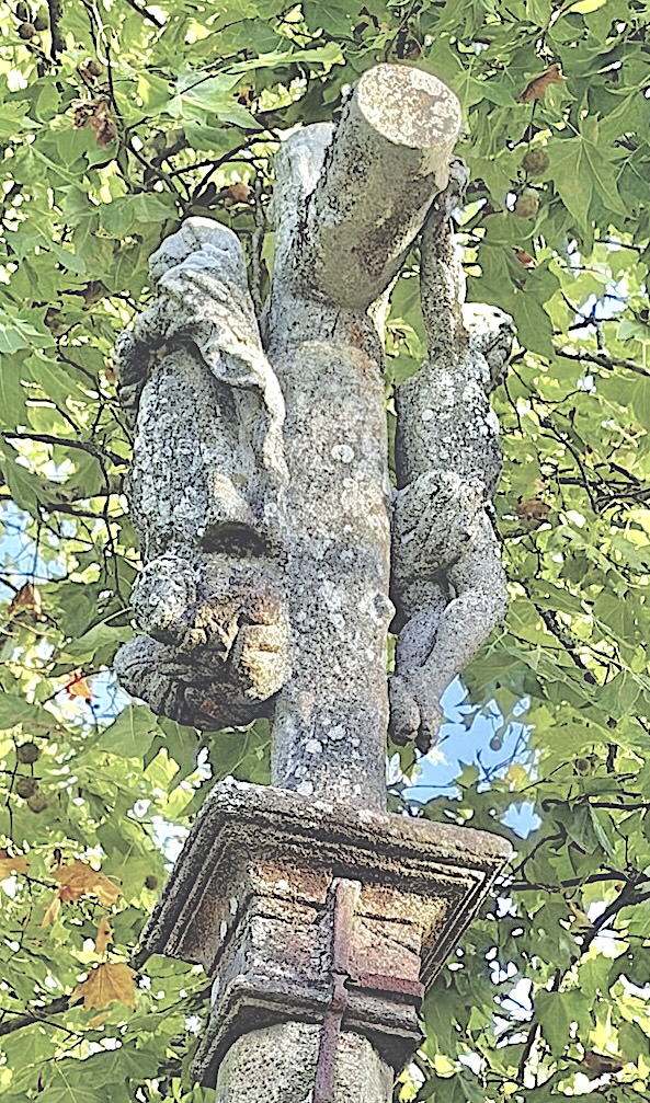 A stone sculpture depicting two figures on a column, surrounded by green leaves and a clear blue sky.