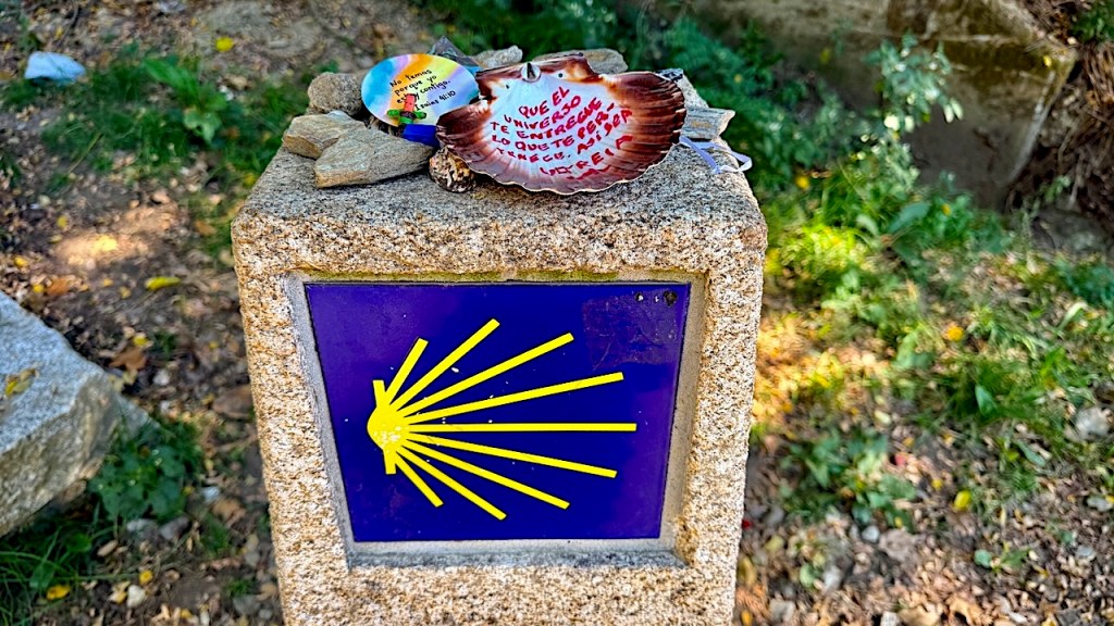 A stone marker featuring a yellow scallop shell design on a blue background, adorned with small colorful stones, and a seashell with handwritten notes.