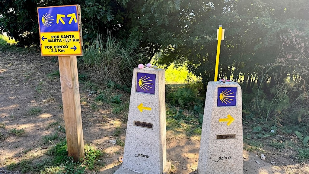 Navigational signpost along a walking path indicating directions to Santa Marta and Conxo, featuring stone markers with yellow arrows and scallop shell symbols.