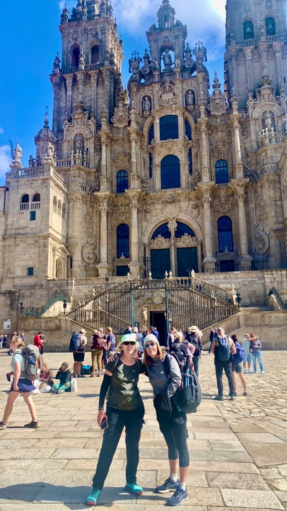 Two women posing in front of a grand, historic building with intricate architecture and tall towers, surrounded by other tourists enjoying a sunny day.