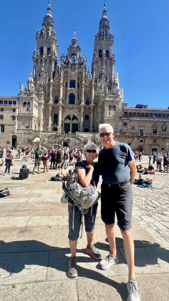 A couple poses for a photo in front of a large, ornate building with intricate architecture in a sunny plaza, surrounded by other visitors.