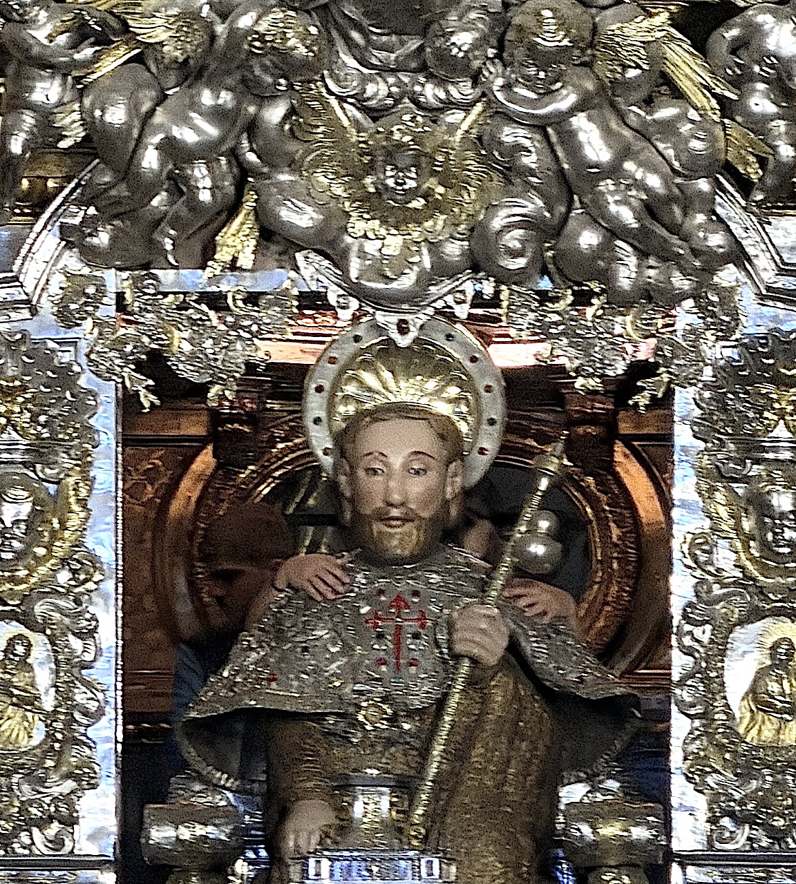 A detailed statue of a religious figure, adorned with a silver robe and a crown, surrounded by decorative angelic figures.
