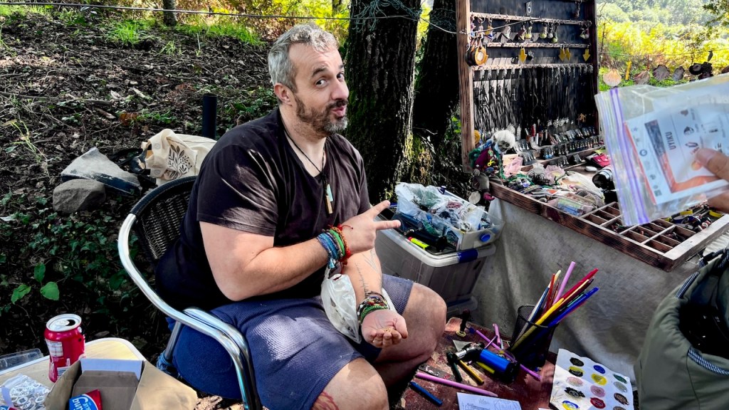 A smiling man sitting on a chair outdoors, sporting bracelets and a casual outfit, gestures peace with one hand while holding paperwork in the other, surrounded by a display of colorful art supplies and various items on a table.