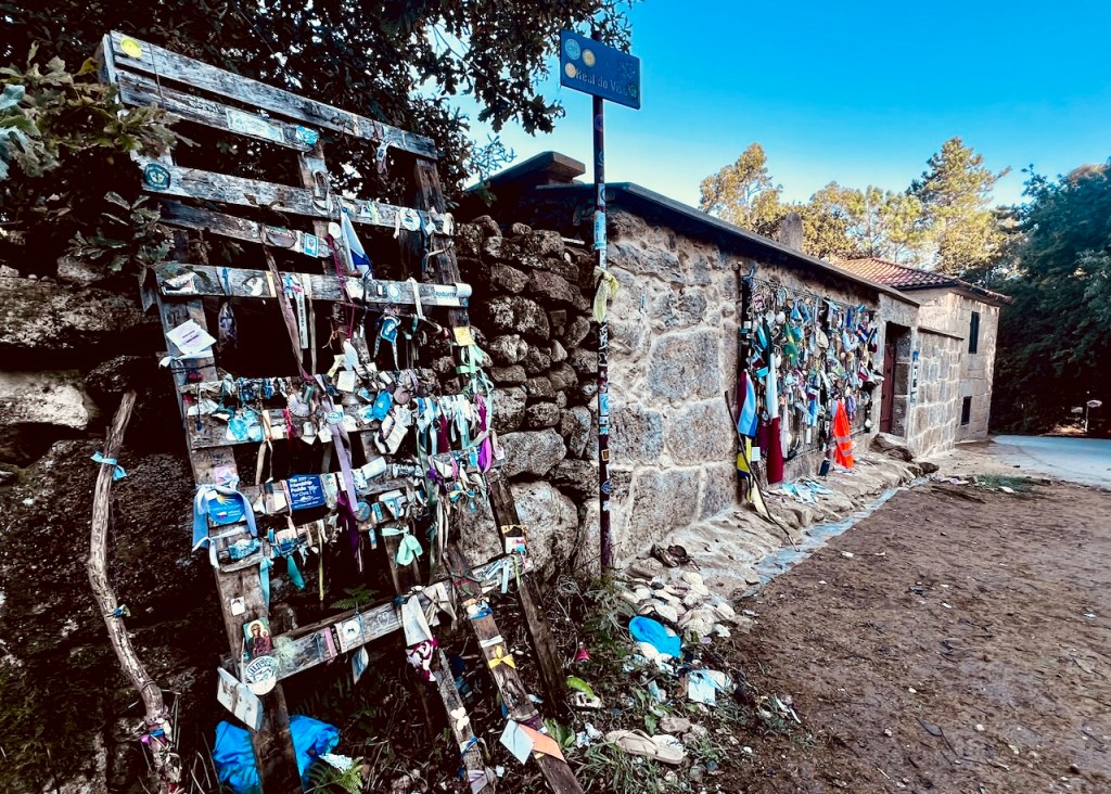A wooden display covered with colorful ribbons and tokens stands against a stone wall near a building, with a street sign visible above.