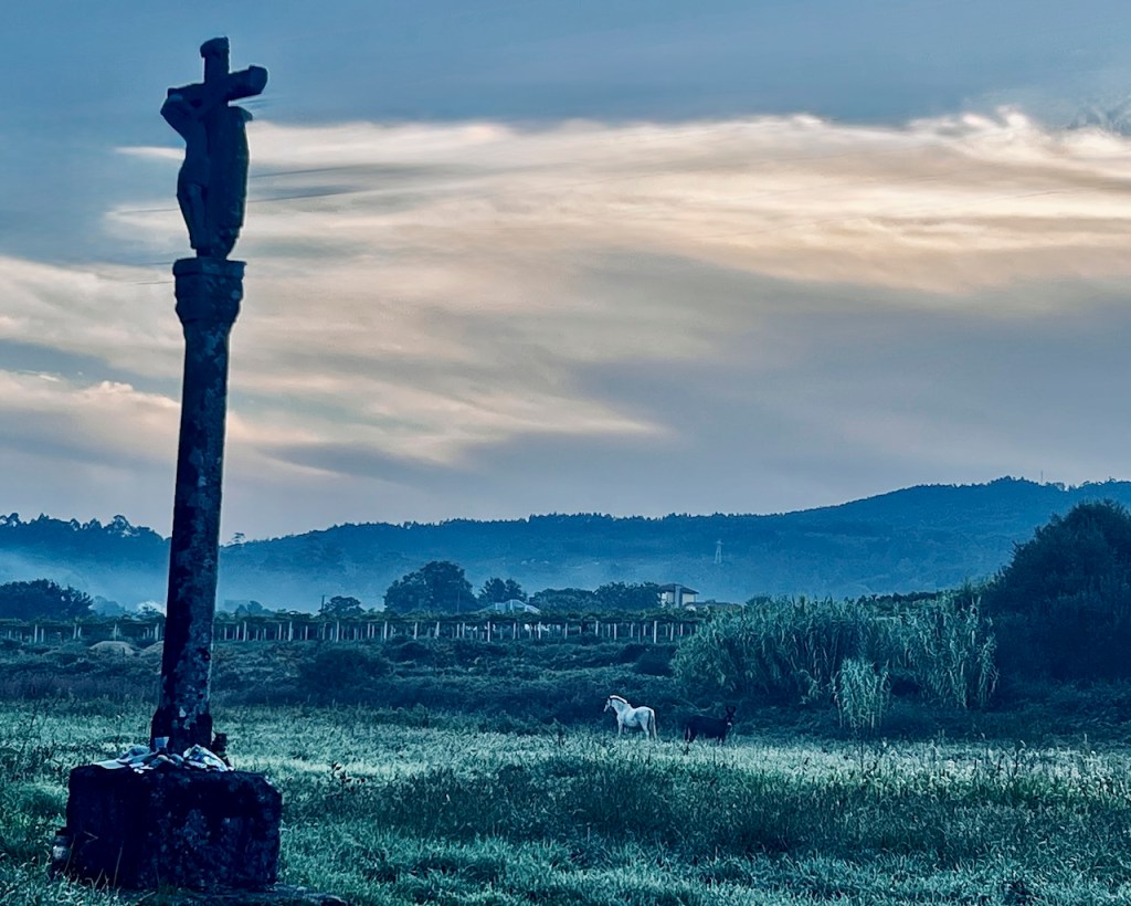 A stone cross statue stands in the foreground with two horses grazing in a misty field, surrounded by lush greenery and rolling hills under a cloudy sky.