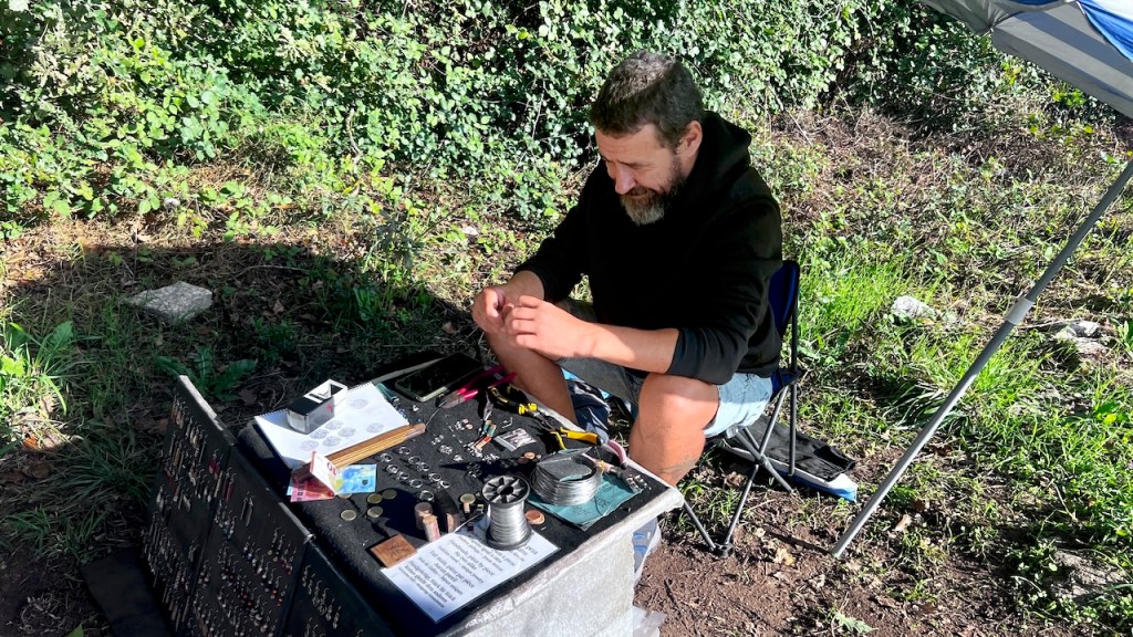 A man sitting at a table outdoors, engaged in a craft or repair activity, surrounded by various tools and materials, with greenery in the background.
