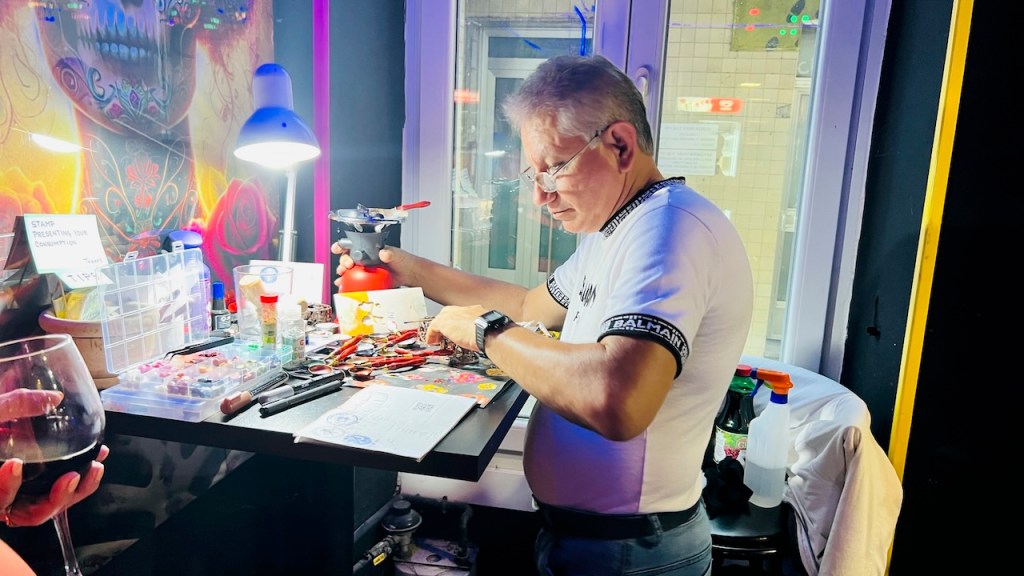 A man working at a cluttered table with various tools and materials, illuminated by a desk lamp, in a colorful room with a decorative mural.