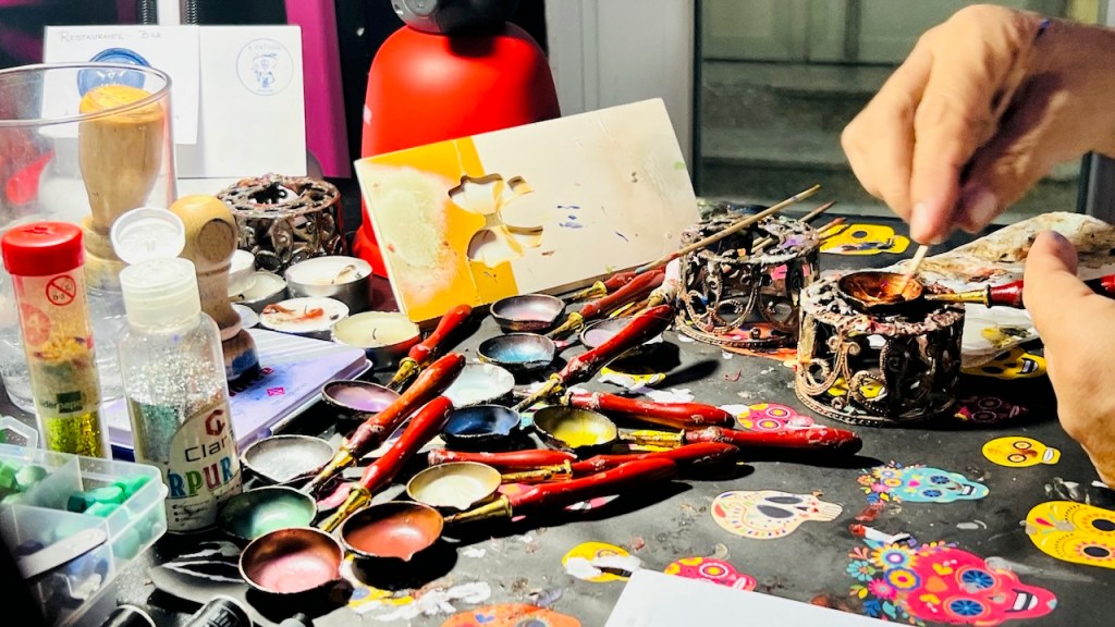 A cluttered artist's workspace filled with paintbrushes, paint palettes, and various art supplies, with a person mixing paint in the foreground.
